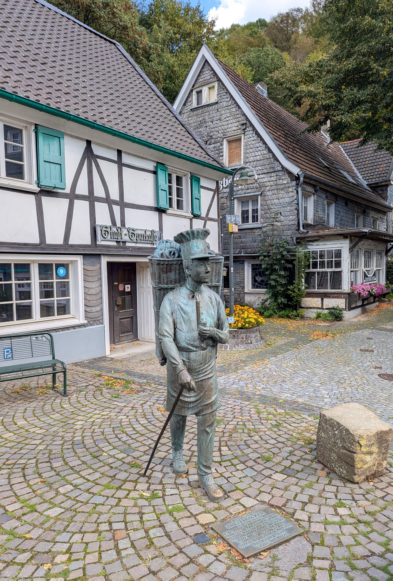 Der Burger Brezelmann, eine Bronzefigur mit Brezelkorb, vor Fachwerk- und Schieferhäusern in Unterburg