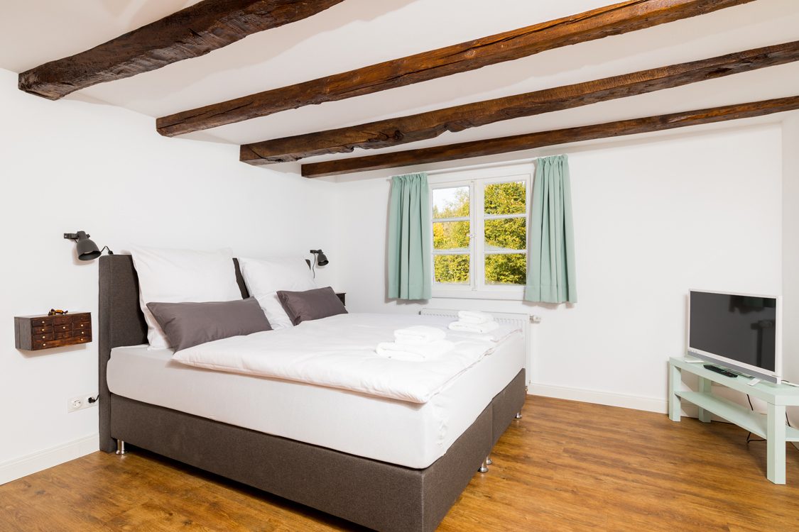 Die Brezelkammer: Doppelbett unter dunklen Holzbalken, Fenster mit mintgrünen Vorhängen